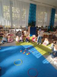 Dzieci siedzą w kole na dywanie. Na środku dywanu leżą klocki w kształcie figur geometrycznych