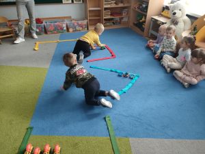 Na dywanie ułożone są różne kształty z szarf- parkingi. Dzieci siedzą na dywanie. Dwóch chłopców parkuje zabawkowe samochodziki w wyznaczonych miejscach