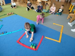 Na dywanie ułożone są różne kształty z szarf- parkingi. Dzieci siedzą na dywanie. Dwóch chłopców parkuje zabawkowe samochodziki w wyznaczonych miejscach