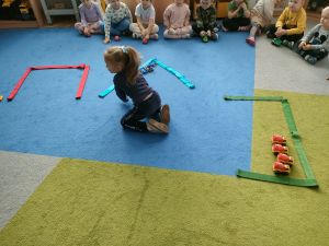 Na dywanie ułożone są różne kształty z szarf- parkingi. Dzieci siedzą na dywanie. Dziewczynka parkuje zabawkowe samochodziki w wyznaczonych miejscach