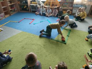 Na dywanie ułożone są różne kształty z szarf- parkingi. Dzieci siedzą na dywanie. Troje dzieci parkuje zabawkowe samochodziki w wyznaczonych miejscach