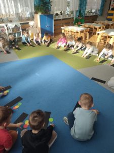 Dzieci siedzą w kole a przed nimi leży prostokątna kartka i kolorowe kółka z papieru