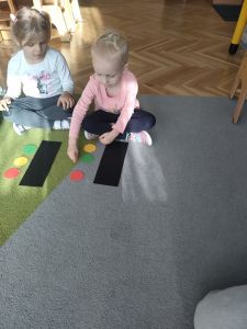 Dwie dziewczynki siedzą na dywanie a przed nimi leży czarna prostokątna kartka i kółka w kolorze żółtym, zielonym i czerwonym