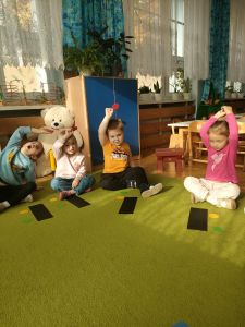 Dzieci siedzą w kole a przed nimi leży prostokątna kartka i kolorowe kółka z papieru