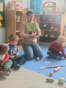 Dzieci siedzą na dywanie, a nauczycielka pokazuje warzywa 