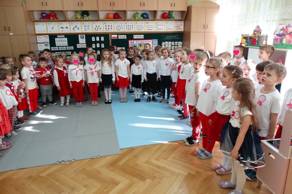 Dzieci z wszystkich grup z przedszkola stoją ustawione w półkole. Dzieci ubrane są na galowo, w biało czerwone stroje. 