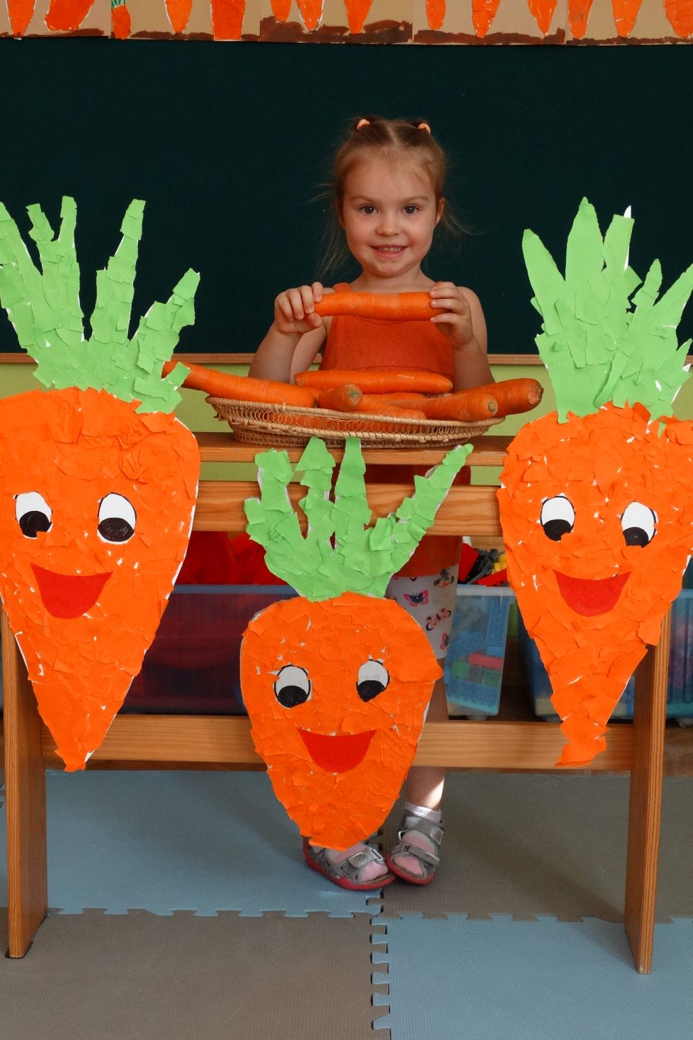 Dziewczynka ubrana na pomarańczowo trzyma w ręce marchewkę, z przed nią stoi półka z marchewkami z papieru