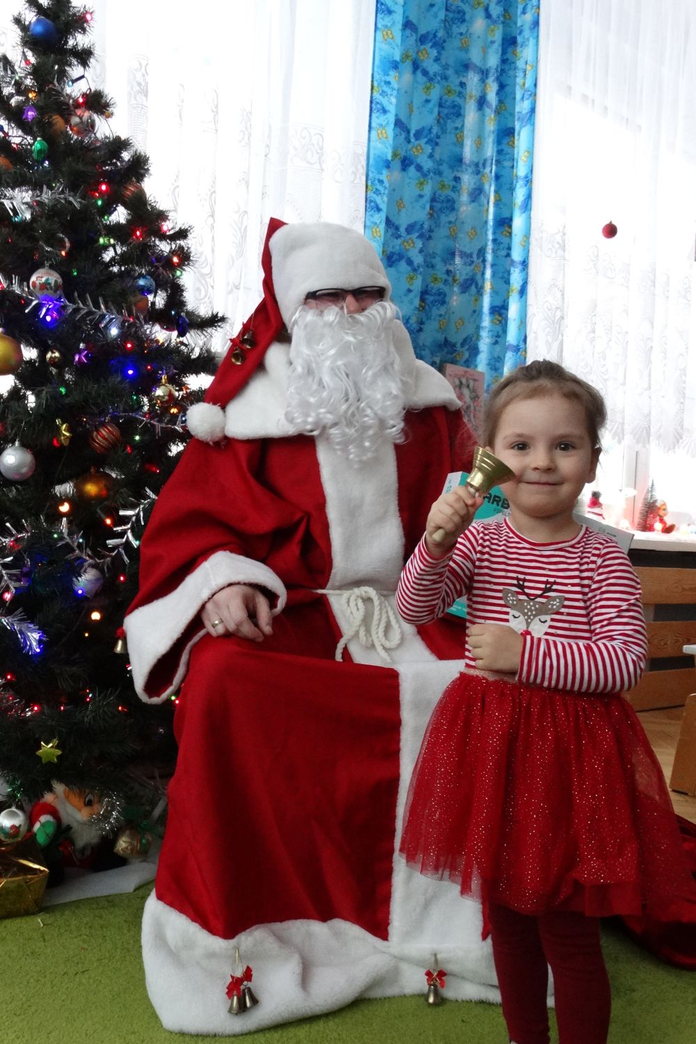 Dziewczynka z grupy 2 stoi przed Mikołajem i trzyma w ręce dzwonek św. Mikołaja. Mikołaj siedzi na krześle obok choinki