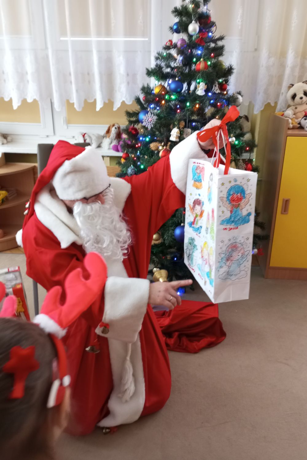 Święty Mikołaj kuca na dywanie, w tle widać choinkę. Mikołaj trzyma w ręce torbę prezentową ozdobioną rysunkami dzieci z grupy 3