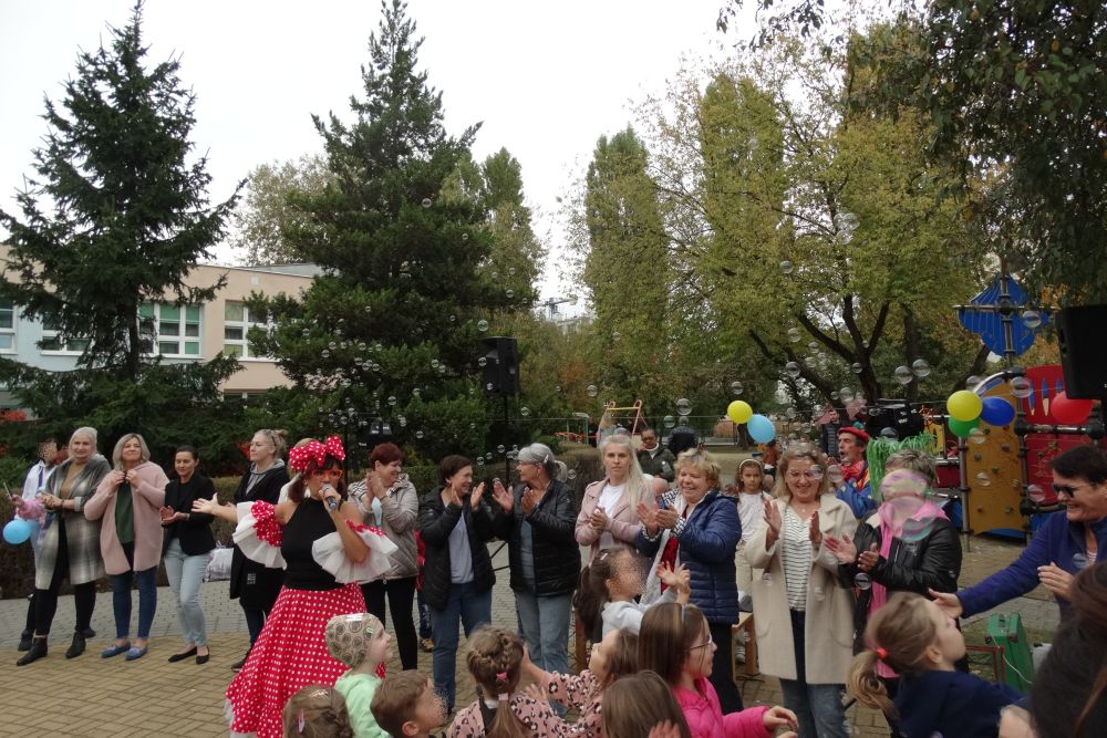 Pracownicy przedszkola stoją w szeregu na placu zabaw a przed nimi stoi prowadząca w stroju myszki mini i dzieci z rodzicami