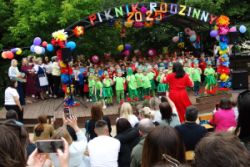 Na scenie dzieci z wszystkich grup śpiewają piosenkę, obok sceny widać stojących nauczycieli. Widać też publiczność