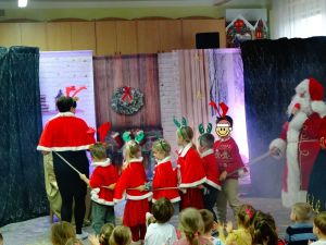 Na scenie jedna z Pań z przedszkola i 6 dzieci ubrana w mikołajowe kubraczki i z reniferowymi opaskami idą po scenie