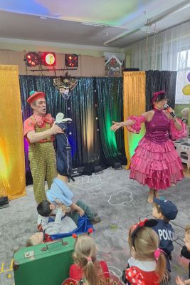 Na scenie prowadząca mówi do mikrofonu. Obok stoi Magik, który przytrzymuje nogi chłopca, który robi figurę akrobatyczną z innym chłopcem.