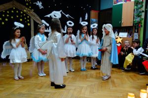 W tle dekoracja świąteczna- na ciemnym materiale stajenka Betlejemska, aniołki i gwiazdy. Dzieci ubrane w stroje pasterzy, aniołków, Maryi, Józefa, Królów i w stroje ludowe. Na środku sceny stoją dzieci przebrane za aniołki, archanioł i Maryja