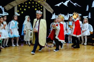 W tle dekoracja świąteczna- na ciemnym materiale stajenka Betlejemska, aniołki i gwiazdy. Dzieci ubrane w stroje pasterzy, aniołków, Maryi, Józefa, Królów i w stroje ludowe. Na środku sceny idą Królowie z darami