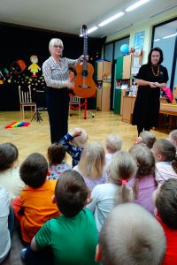 Dzieci siedzą na dywanie. Pani gitarzystka pokazuje gitarę, obok stoi Pani od rytmiki