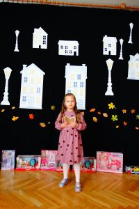 Dziewczynka stoi na środku sceny i mówi do mikrofonu. W ręce trzyma piernikowe serduszko. W tle dekoracja przedstawiające białe budynki.