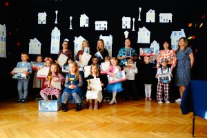 Zdjęcie grupowe wszystkich osób biorących udział w konkursie. W ręce trzyma piernikowe serduszko. W tle dekoracja przedstawiające białe budynki.