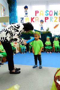 Pani dyrektor pasuje na przedszkolaka chłopca, kładąc na jego ramieniu duży ołówek