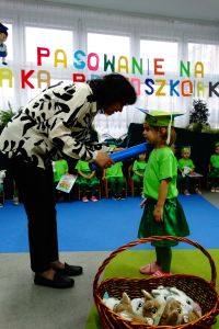 Pani dyrektor pasuje na przedszkolaka dziewczynkę, kładąc na jej ramieniu duży ołówek