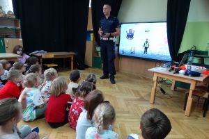 policjant pokazuje dzieciom prezentację na tablicy multimedialnej. Dzieci siedzą na dywanie i uważnie słuchają