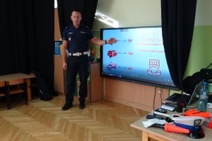 policjant pokazuje dzieciom prezentację na tablicy multimedialnej. Policjant opowiada o numerach alarmowych