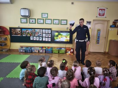 Dzieci siedzą na podłodze, przed nimi stoi policjant i pokazuje policyjną czapkę. W tle włączony telewizor z filmikiem ze spongeBob