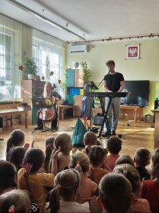Dzieci siedzą i słuchają koncertu, Na scenie Pan grający na kibordzie i Pani grająca na wiolonczeli