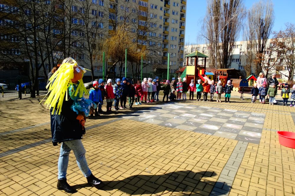 Na zdjęciu dzieci z wszystkich grup stoją na placu przedszkolnym. Jedna z Pań trzyma kolorową marzannę.