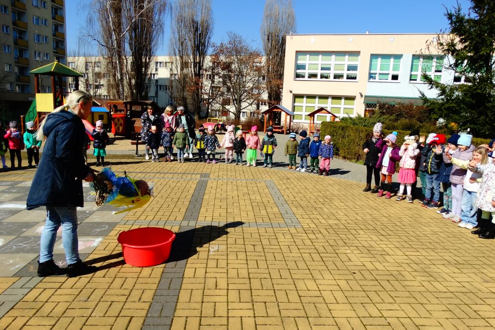 Dzieci ze wszystkich grup stoją na placu przedszkolnym. Jedna z Pań trzyma marzannę nad miską z wodą.