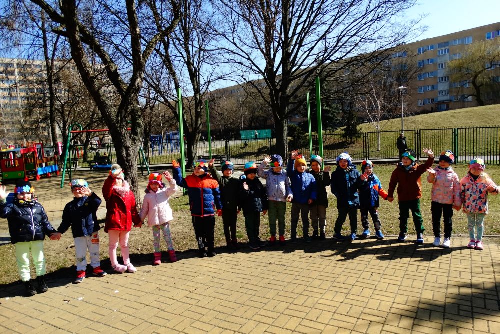 Dzieci z grupy 4 stoją na dworze na placu przedszkolnym, na czapkach mają opaski z kwiatkami. Dzieci machają.