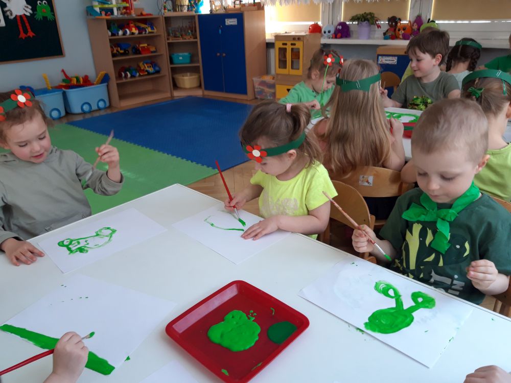 Dzieci z grupy 2, siedzą przy stoliku i malują obrazki zieloną farbą. Dzieci ubrane się na zielono. Dziewczynki mają zielone papierowe opaski na głowach ozdobione kwiatkami.