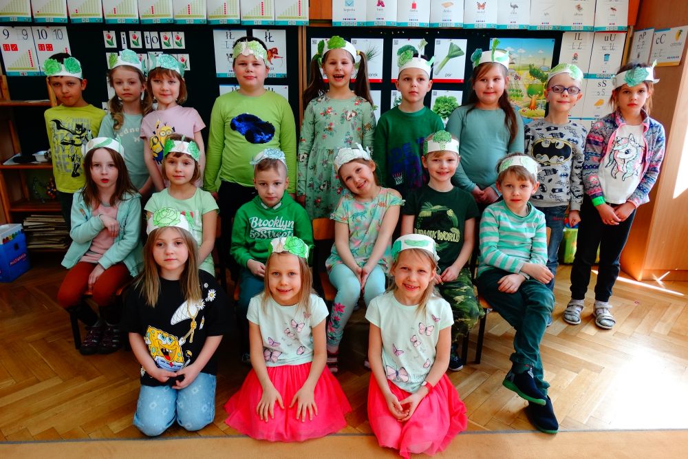 Zdjęcie grupowe dzieci z grupy 5. Dzieci mają ubrania w kolorze zielonym. Na głowach mają papierowe opaski z naklejonymi zielonymi warzywami.