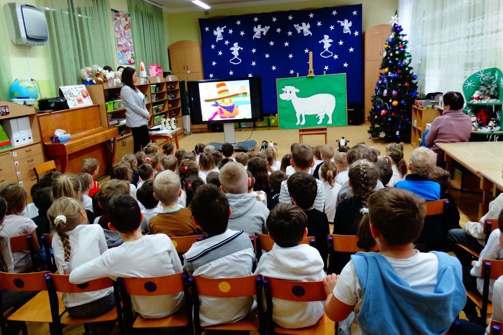 Na zdjęciu dzieci z całego przedszkola siedzą tyłem, patrzą na ekran tablicy multimedialnej na której wyświetlana jest bajka.