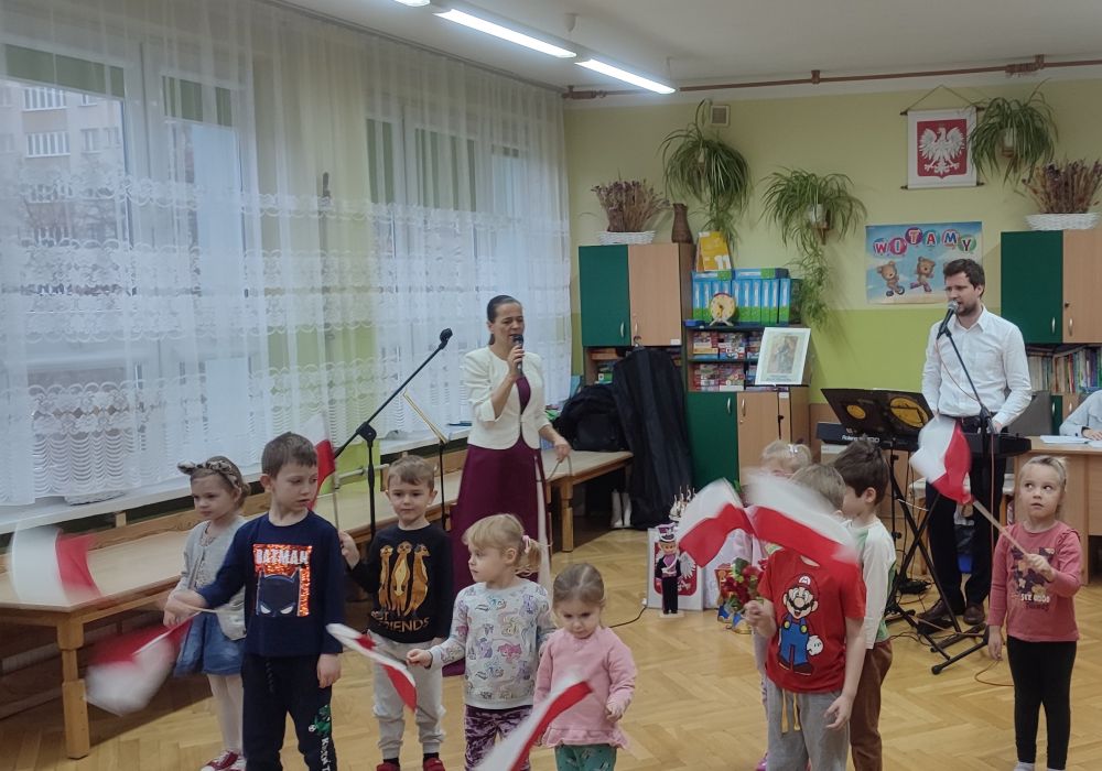 Kilkoro dzieci stoi podczas występu machając flagami. Z tyłu stoi Pani ubrana w biało czerwoną sukienkę i Pan grający na kibordzie