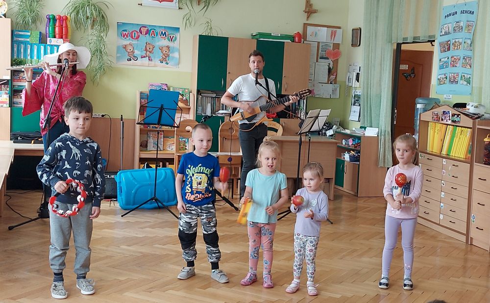 5 dzieci stoi i gra na instrumentach. Za nimi stoi pani i gra na flecie poprzecznym i pan gra na gitarze 