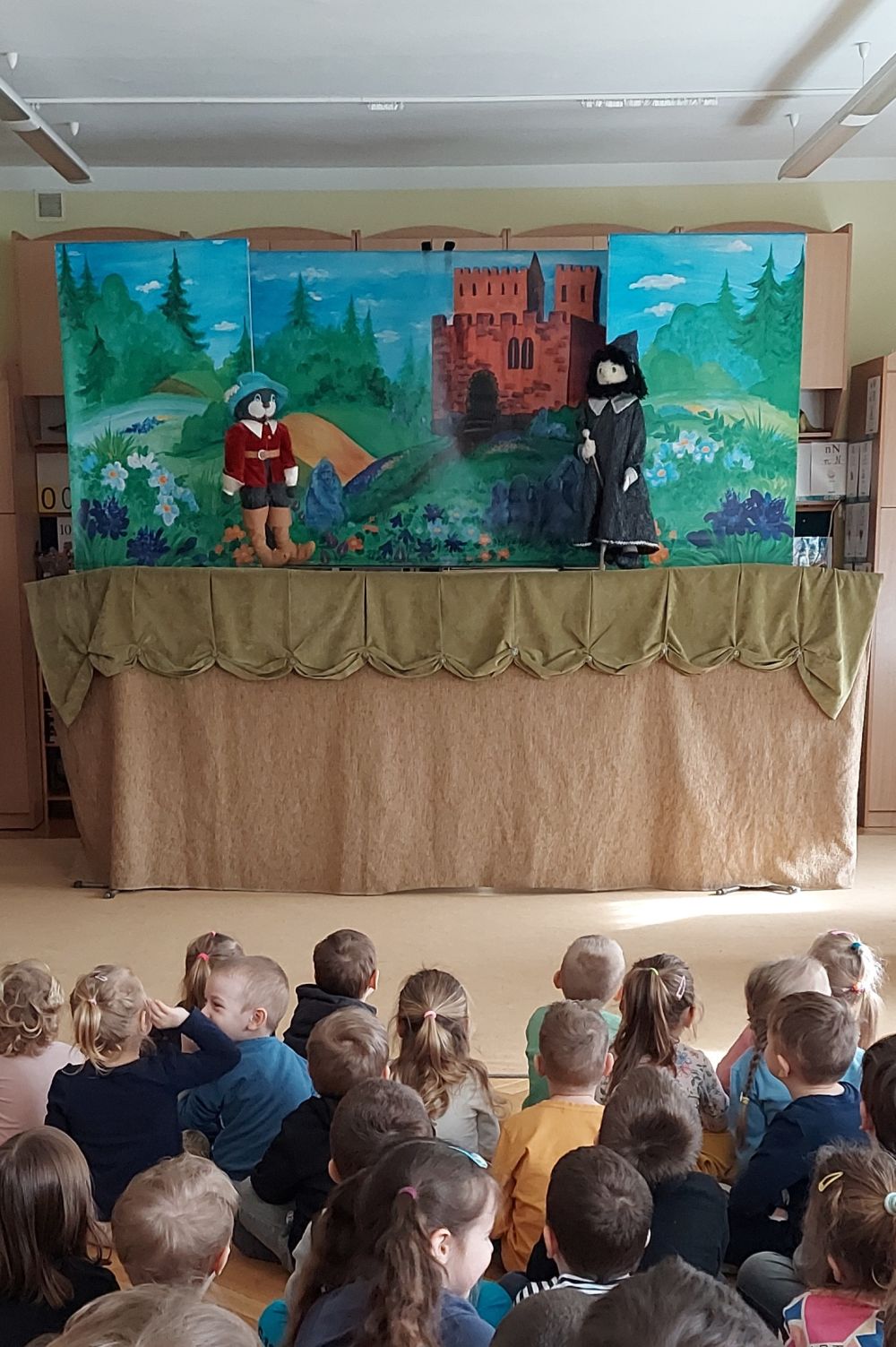 Dzieci siedzą na dywanie i patrzą na teatrzyk kukiełkowy. Widać dwie kukiełki kota i Janka. w tle namalowany krajobraz i zamek