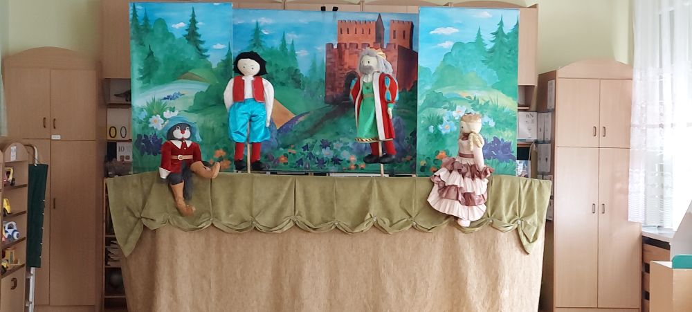 Dzieci siedzą na dywanie i patrzą na teatrzyk kukiełkowy. Widać cztery kukiełki kota, Janka, króla i księżniczkę. w tle namalowany krajobraz i zamek