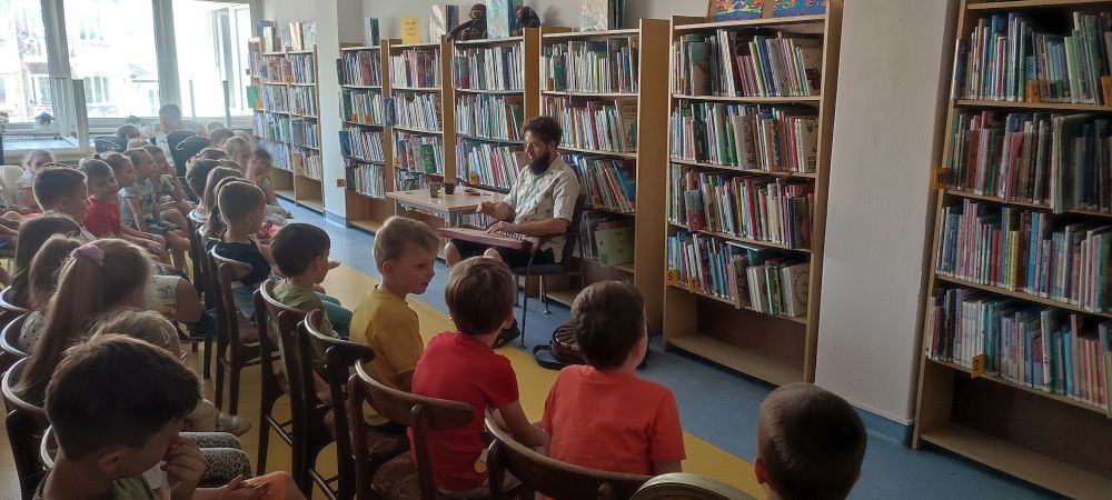 Dzieci siedzą na krzesłach w bibliotece przodem do Pana grającego na instrumencie