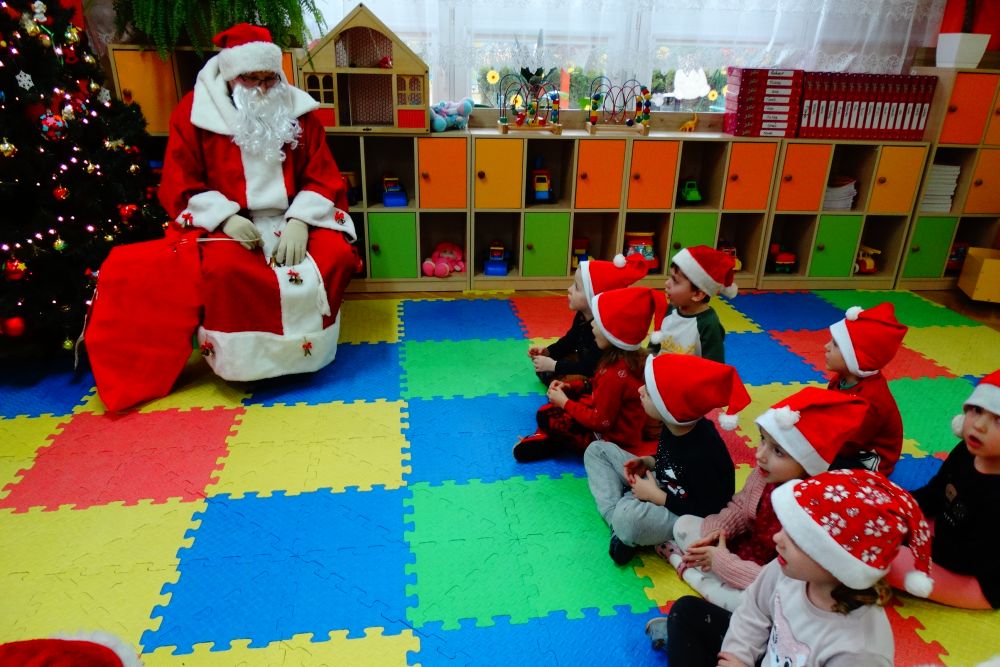 Dzieci siedzą na dywanie w sali, patrząc na Mikołaja, który siedzi na krzesełku obok choinki