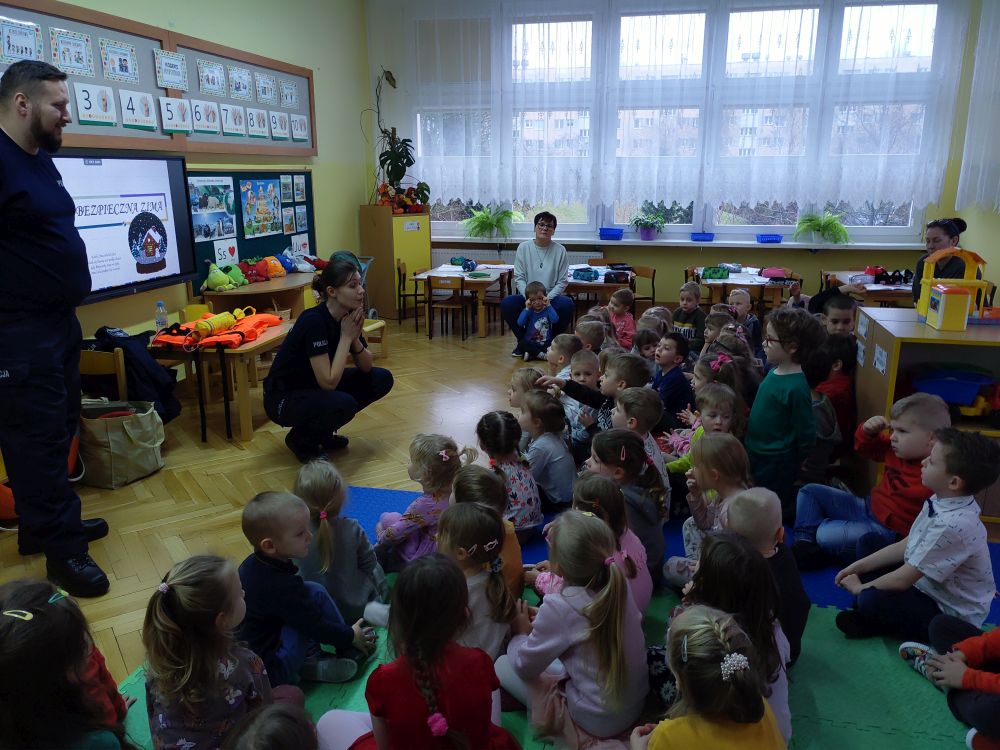 Dzieci z przedszkola siedzą na dywanie patrząc na ekran na którym wyświetlana jest prezentacja. Obok ekranu stoją funkcjonariusze policji rzecznej- kobieta i mężczyzna