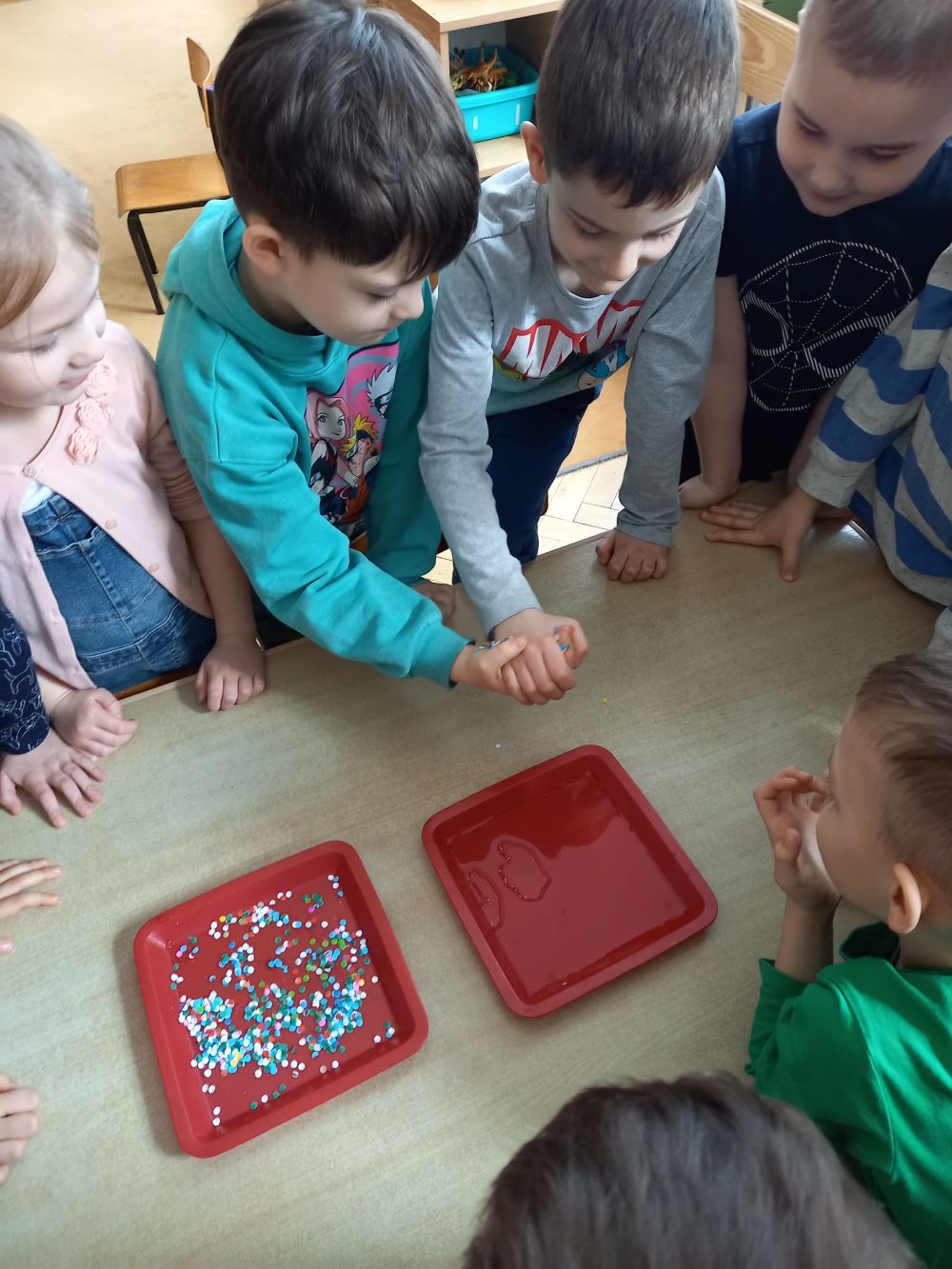 Kilkoro dzieci stoi przy stoliku. Na stoliku leżą dwie czerwone tacki- z wodą oraz konfetti. Dwóch chłopców podaje sobie ręce. Jeden ma rękę oblepioną konfetti. 