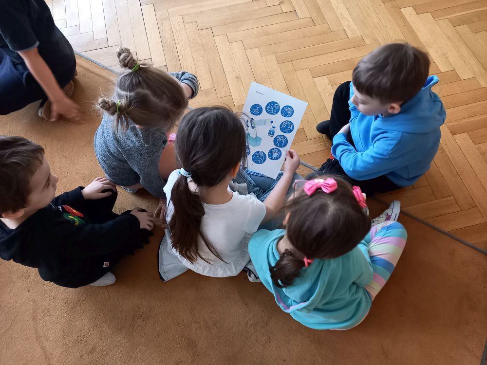 Dzieci siedzą na dywanie i oglądają kartkę z obrazkami jak należy prawidłowo myć ręce