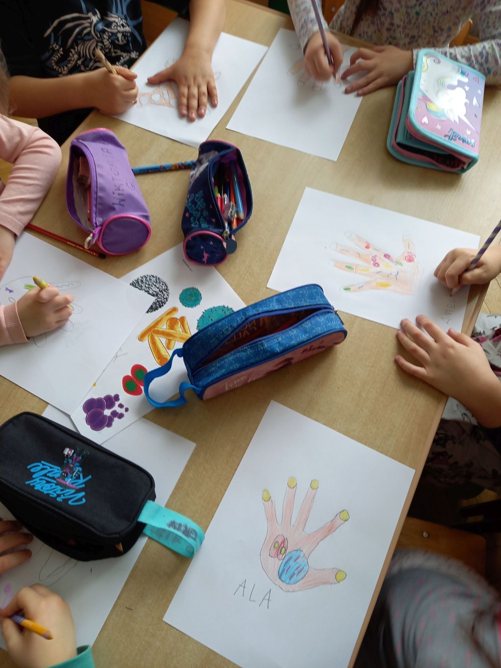 dzieci siedzą przy stoliku i rysują bakterie na odrysowanych dłoniach na kartach papieru
