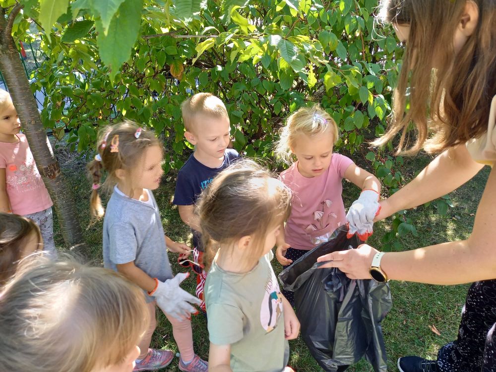 Nauczycielka trzyma worek na śmieci a dzieci wrzucają do niego zebrane śmieci