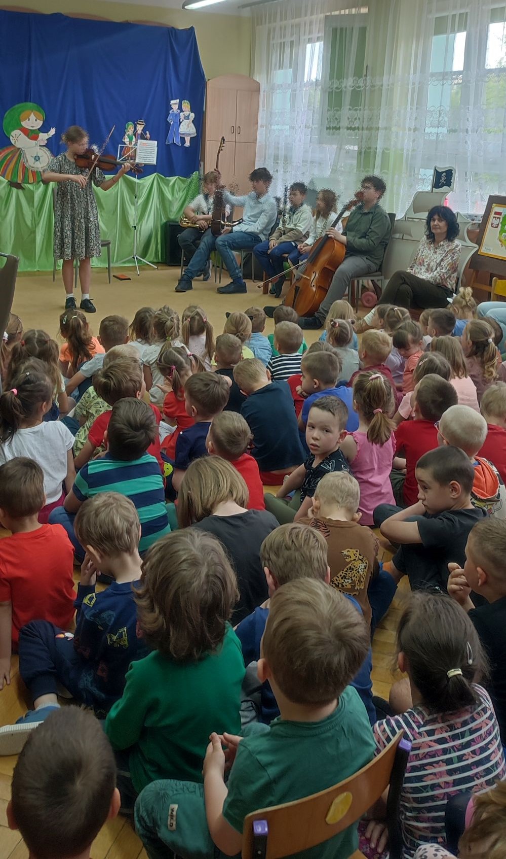 dzieci siedzą na dywanie i patrzą na dziewczynkę grającą na skrzypcach