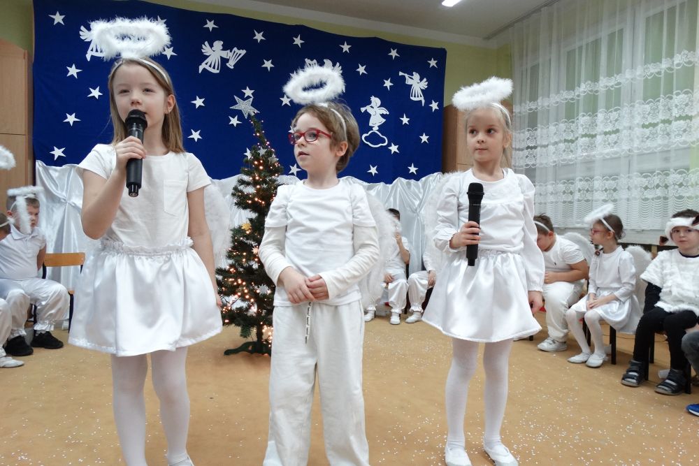 Troje dzieci w strojach aniołków stoi na środku i mówi do mikrofonu. Reszta dzieci siedzi z tyłu w półkolu