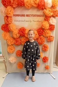 Dziewczynka stoi a w tle dekoracja kwiatowa z napisem Konkurs recytatorski