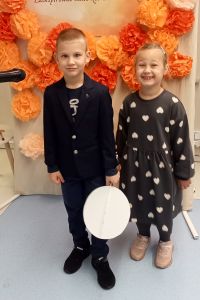 Dziewczynka i chłopiec stoją a w tle dekoracja kwiatowa z napisem Konkurs recytatorski