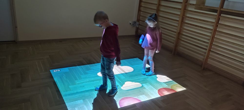 Chłopiec i dziewczynka bawią się na magicznym dywanie, nadeptując na wyświetlane balony.